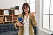 © Krakenimages.com - Young brunette woman working at the office with smartphone pointing thumb up to the side smiling happy with open mouth
