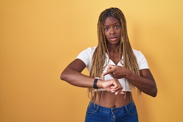 Wall Mural - African american woman with braided hair standing over yellow background in hurry pointing to watch time, impatience, upset and angry for deadline delay