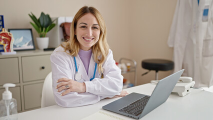 Wall Mural - Young beautiful hispanic woman doctor using laptop working sitting on table with arms crossed gesture at clinic