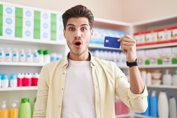 Sticker - Young hispanic man at pharmacy drugstore holding credit card scared and amazed with open mouth for surprise, disbelief face