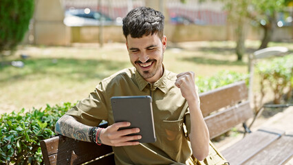 Sticker - Young hispanic man tourist using touchpad sitting on bench celebrating at park