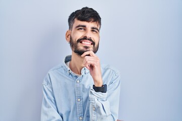Wall Mural - Young hispanic man with beard standing over blue background with hand on chin thinking about question, pensive expression. smiling and thoughtful face. doubt concept.