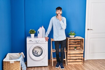 Poster - Young hispanic man with beard doing laundry standing at laundry room covering mouth with hand, shocked and afraid for mistake. surprised expression