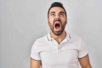 Sticker - Young hispanic man with beard wearing casual clothes over white background angry and mad screaming frustrated and furious, shouting with anger. rage and aggressive concept.