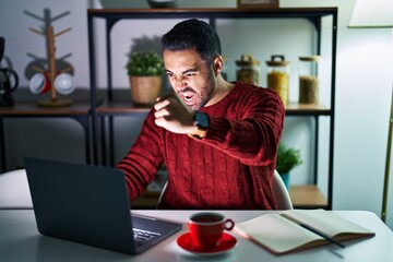 Wall Mural - Young hispanic man with beard using computer laptop at night at home angry and mad raising fist frustrated and furious while shouting with anger. rage and aggressive concept.