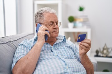 Canvas Print - Middle age grey-haired man talking on smartphone holding credit card sitting on sofa at home