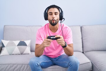 Poster - Hispanic young man playing video game holding controller sitting on the sofa making fish face with lips, crazy and comical gesture. funny expression.