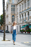 Fototapeta Na drzwi - Smiling woman wallking on historic street Batumi city in Georgia. Young woman wearing jacket and jeans holding bag. In front of architectural buildings. Vertical composition. 