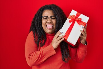 Poster - Plus size hispanic woman holding gift sticking tongue out happy with funny expression.