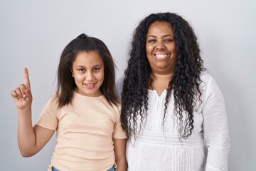 Sticker - Mother and young daughter standing over white background showing and pointing up with finger number one while smiling confident and happy.