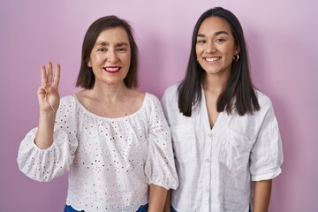 Wall Mural - Hispanic mother and daughter together showing and pointing up with fingers number three while smiling confident and happy.