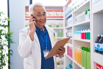 Canvas Print - Middle age grey-haired man pharmacist talking on smartphone reading document at pharmacy