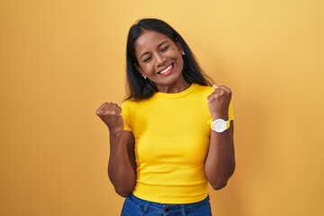 Young indian woman standing over yellow background very happy and excited doing winner gesture with arms raised, smiling and screaming for success. celebration concept.