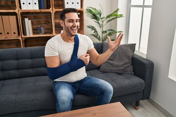 Sticker - Young hispanic man with beard wearing arm on sling sitting at therapy consult celebrating victory with happy smile and winner expression with raised hands