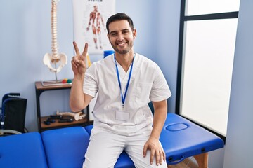 Wall Mural - Young hispanic man with beard working at pain recovery clinic showing and pointing up with fingers number two while smiling confident and happy.