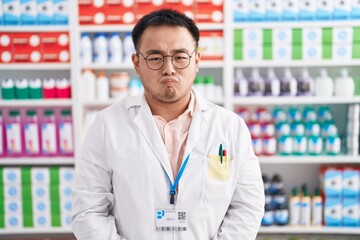 Poster - Chinese young man working at pharmacy drugstore puffing cheeks with funny face. mouth inflated with air, crazy expression.