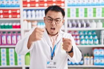 Poster - Chinese young man working at pharmacy drugstore angry and mad raising fists frustrated and furious while shouting with anger. rage and aggressive concept.