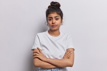 Studio Shot Of Serious Indian Woman With Dark Hair Combed In Bun Keeps Arms Folded Waits For Explanations Listens Attentively Information Dressed In Basic T Shirt Isolated Over White Background.
