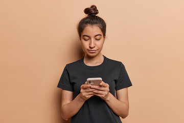 Photo of serious dark haired latin woman concentrated in smartphone chatting on internet working or studying online watches video dressed in casual black t shirt isolated over brown background