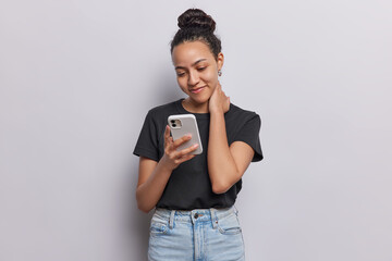 Wall Mural - Pleased young Latin woman glances at her smartphone engrossed in checking sms has focused gaze and faint glow of screen smiles gently dressed in casual black t shirt and jeans keeps hand on neck