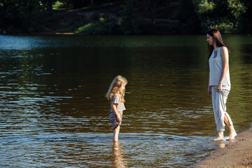 A five-year-old cheerful girl and her mother are walking by the lake, barefoot in the water, having fun on a summer day.