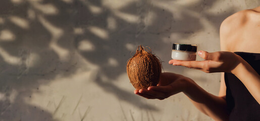 Close-up of woman hands holding coconut cream for skin at textured wall backdrop. Arms of young lady with cosmetic cream in shadow sunlight. Concept of cosmetology and self care. Copy ad text space