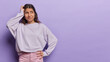 © WHstudio Leushin N - Horizontal shot of confused Latin woman scratches head focused aside tries to make decision wears casual pullover and pink trousers isolated over purple background copy space for your advertisement