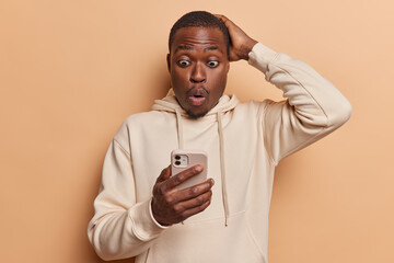 Stupefied young African man checks newsfeed in social networks on cell phone finds out shocking news keeps hand on head dressed in casual sweatshirt isolated over brown background. Omg concept