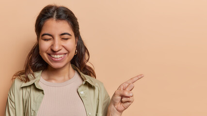 Wall Mural - Horizontal shot of dark haired young Latin woman smiles pleasantly points index finger on copy space shows advertising promo deal keeps eyes closed wears shirt isolated over brown background