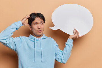 Wall Mural - Thoughtful young Chinese man dressed in casual sweatshirt scratches his head while contemplating perfect advertisement to fill empty speech bubble isolated over brown background. Idea concept