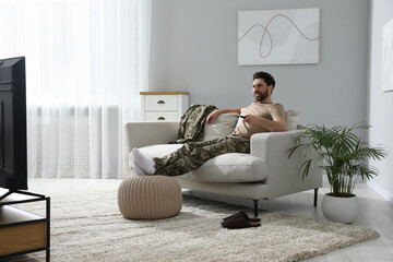 Wall Mural - Happy soldier watching TV on sofa in living room. Military service