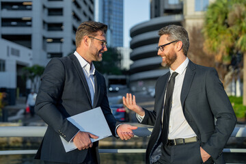 Wall Mural - Business communication. Two business man in suit talking about finance news standing in modern city. Businessmen walking and talking outdoor in New York city. Confident employee talk bysiness idea.