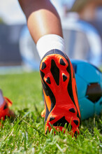 Close-up Of A Girl's Foot With Soccer Boots On The Soccer Field. Blue Ball And Leg Of A Girl With Orange Soccer Boot On The Soccer Field.