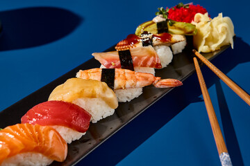 Canvas Print - Macro shot of a sushi set with salmon, tuna, shrimp, eel, and avocado on a black plate. Set against a deep blue background. Modern, minimalist, monochrome setup