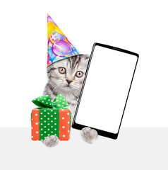 Poster - Cute kitten wearing party cap holds gift box and shows big smartphone with blank screen above empty white banner. isolated on white background