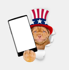Poster - Happy Mastiff puppy wearing like Uncle Sam looking through a hole in paper and showing big smartphone with white blank screen. isolated on white background