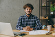 © ViDi Studio - Successful confident happy smart employee business Indian man he wearing casual blue checkered shirt look camera write in notebook read papers sit work at office desk with laptop pc computer indoors.