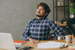 © ViDi Studio - Sad sick ill tired exhausted employee business Indian man he wearing casual blue checkered shirt hold back suffer from pain close eyes sitting working at office desk with laptop pc computer indoors