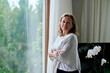 © Westend61 - Thoughtful senior woman standing with arms crossed by window at home