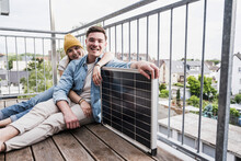 Happy Young Couple Sitting With Solar Panel By Railing On Balcony