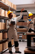 Woman storehouse operator working with digital tablet in warehouse and taking parcel from shelf. African american delivery service storage employee pointing at cardboard boxes