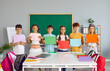© Studio Romantic - Positive elementary school teens boys and girls in casual clothes show colored paper sheets to create appliques or text stand between tables and board. Primary education and back to school concept