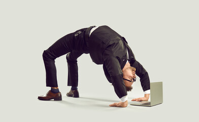 Extravagant curious businessman standing in bridge position looking at laptop screen on gray background. Crazy young man in suit stands on his hands and feet in yoga pose bridge near laptop.