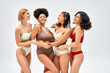 © LIGHTFIELD STUDIOS - Cheerful multiethnic women in colorful lingerie hugging african american friend while standing and posing together isolated on grey, different body types and self-acceptance concept