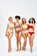 © LIGHTFIELD STUDIOS - Full length of joyful multiethnic women in colorful bras and panties hugging while standing together on grey background, different body types and self-acceptance concept, multicultural models