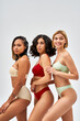 © LIGHTFIELD STUDIOS - Smiling blonde woman in lingerie looking away while standing near multiethnic friends and posing isolated on grey, different body types and self-acceptance concept, multicultural models