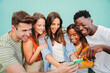 Multiracial group of young friends enjoying and smiling using their mobile phone app at teal blue wall background. Diverse teenage student people having fun watching content on the social media. High