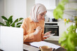 © Johnér - Smiling woman with hijab using cell phone during work from home in kitchen