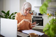 © Johnér - Smiling woman with hijab using cell phone during work from home in kitchen
