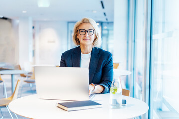 Portrait of 50's confident mature businesswoman looking at camera, middle-aged experienced senior female professional working on laptop in open space office. Female entrepreneur working remotely